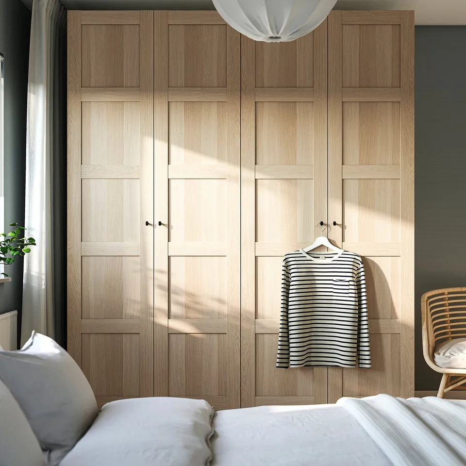 Natural wood PAX wardrobe, tall and narrow beside white bed, hanging shirt, large overhead light.