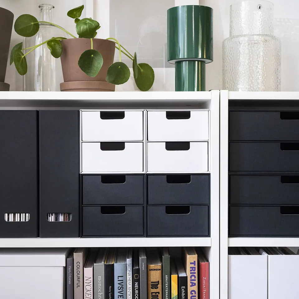 Grey FJÄDERHARV magazine holders with books on white shelves, topped with plants and vases.