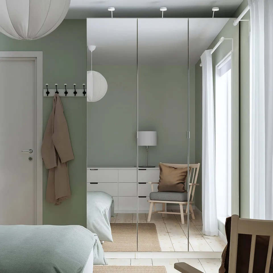 Minimalist bedroom with green walls, white furniture, and a large mirror wardrobe. Hanging coats, bedside lamp, and calm decor.