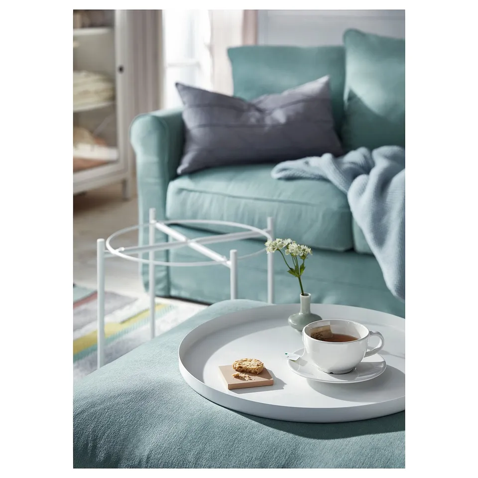 A cosy teal sofa with a round tray on it holds a coffee cup, treat, and small flower vase. The tray rests on a metal stand.