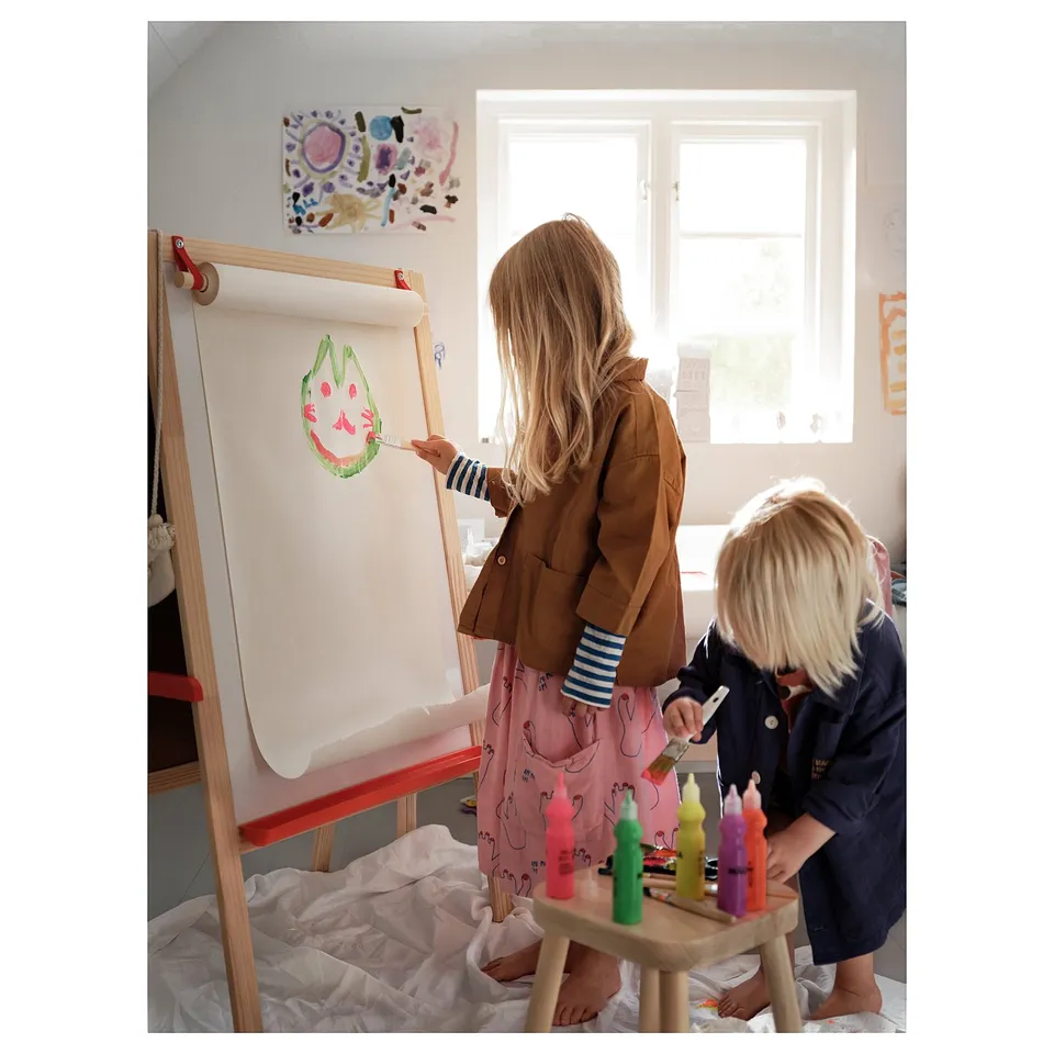 Two children painting on an easel with MÅLA brushes; one wears a brown jacket, the other navy. Colourful artwork and bottles of paint nearby.