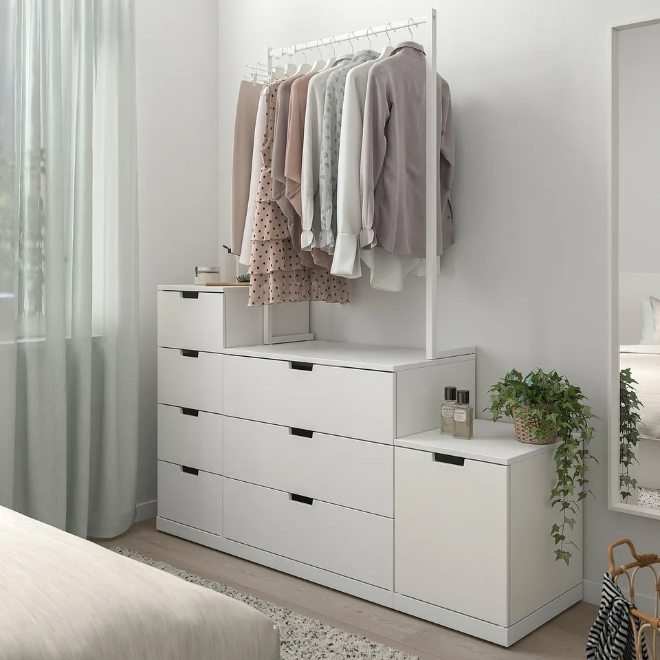 Modern white NORDLI chest with clothes rail, drawers, and plant. Large, rectangular, with light wood floors.
