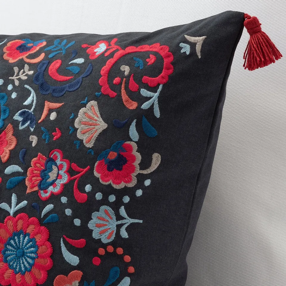 Embroidered cushion with vibrant flowers on dark fabric, featuring a red tassel corner. Detailed, high-quality craftsmanship.