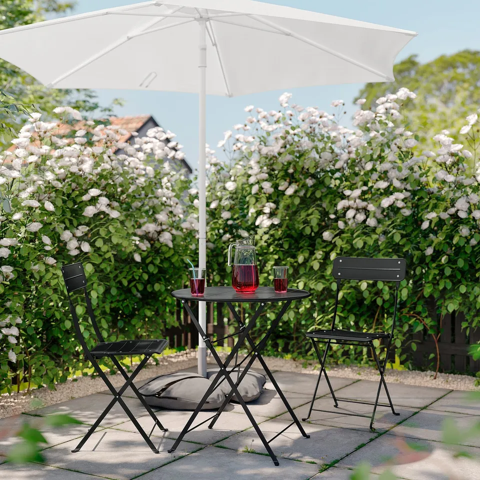 Peaceful garden setting with black SUNDSÖ metal table and chairs, white umbrella, red drinks, surrounded by blooming bushes.
