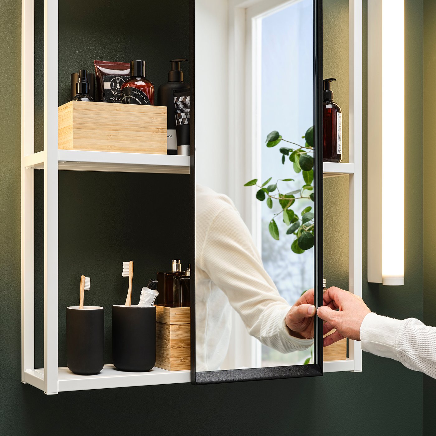 A person adjusts a sliding bathroom mirror, revealing shelving with toiletries and a window view.