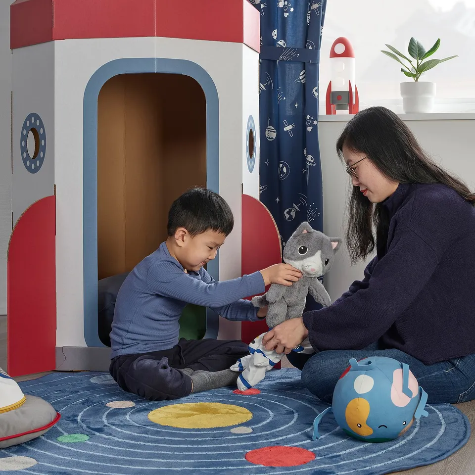 Child plays with cat toy, guided by person, on space rug beside playhouse.