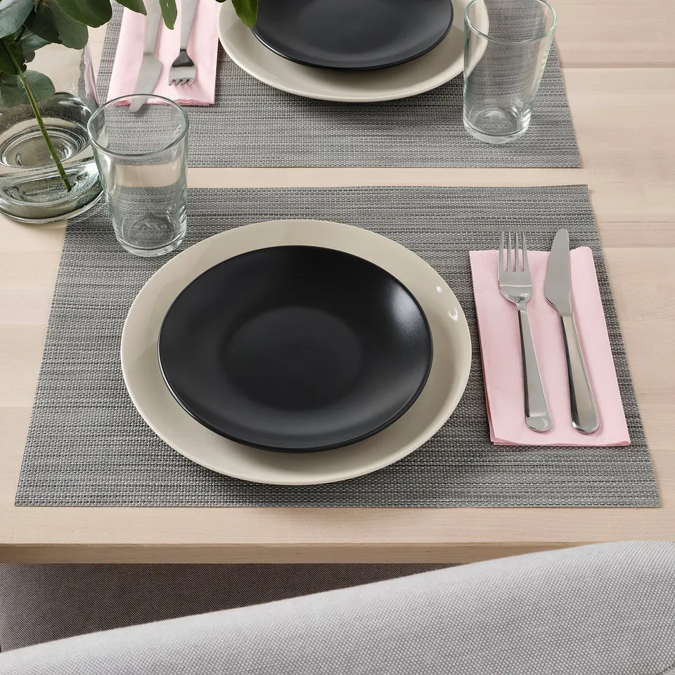 Two place settings on a table with grey placemats, black and white plates, pink napkins, forks, knives, glasses, and a small candle.