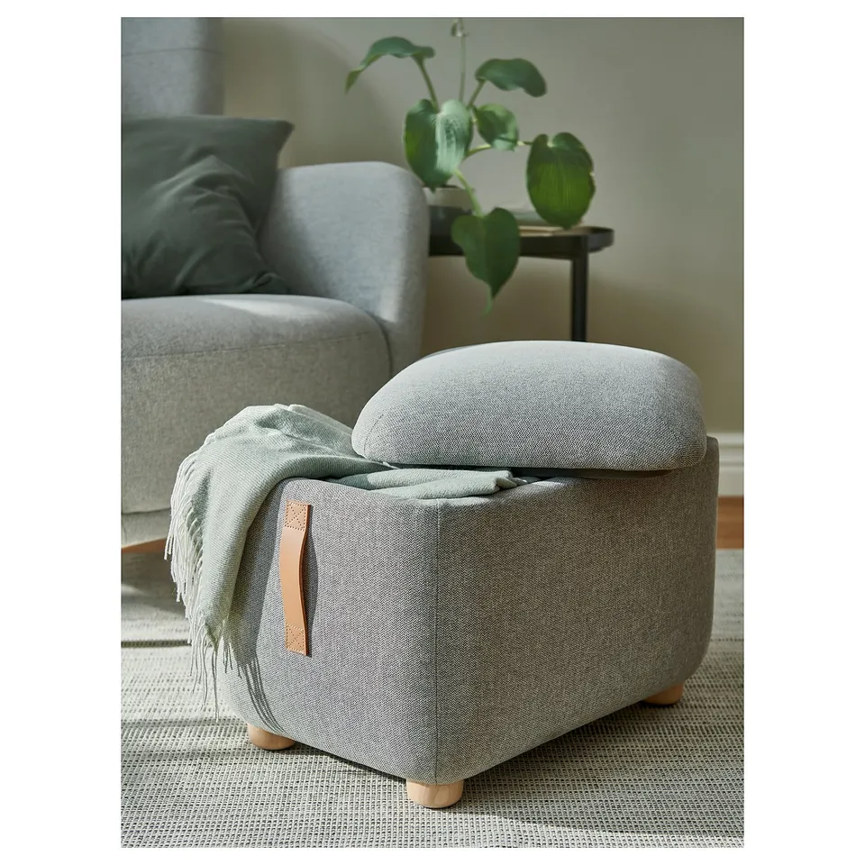 Grey ososhman footstool with leather handle & wooden legs; small, near chair & plant.