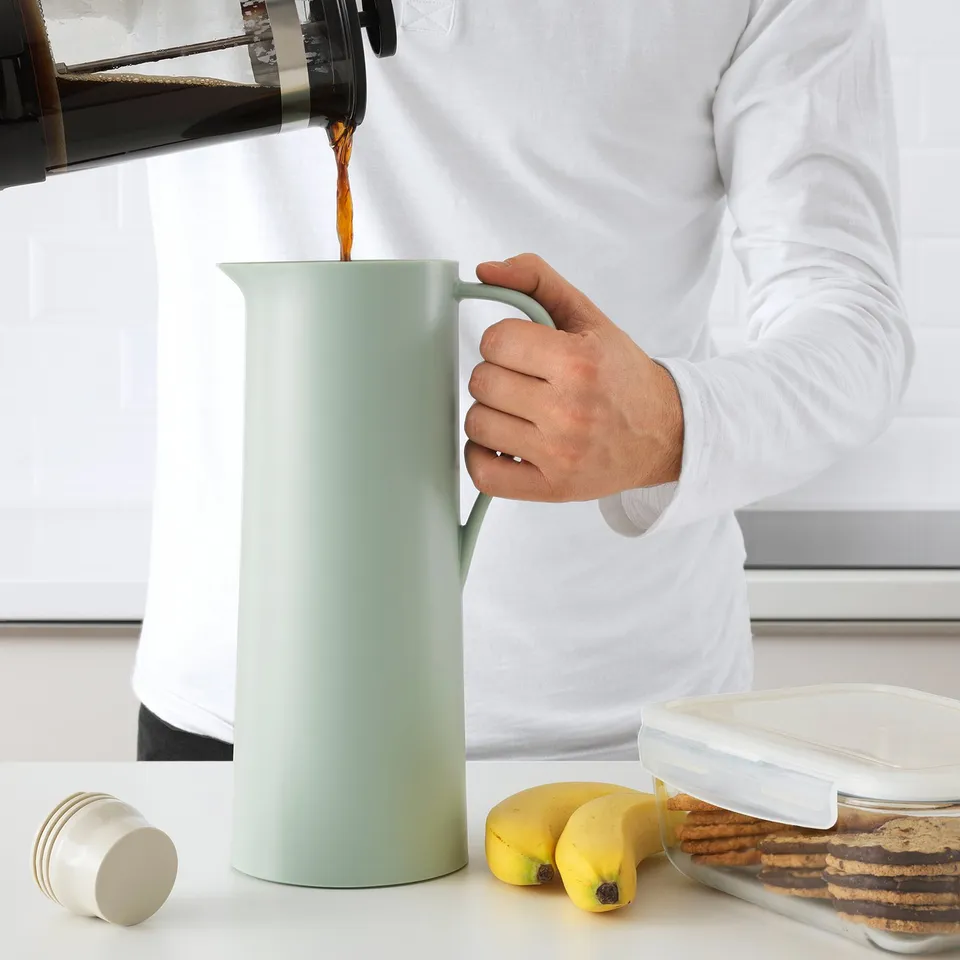 Person pours coffee into green insulated flask with partial lid. Table has bananas and cookies.