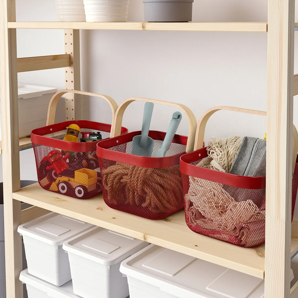 Three red mesh baskets with wooden handles on a wooden shelf, storing toys and fabric items; under them, white plastic containers on another shelf.