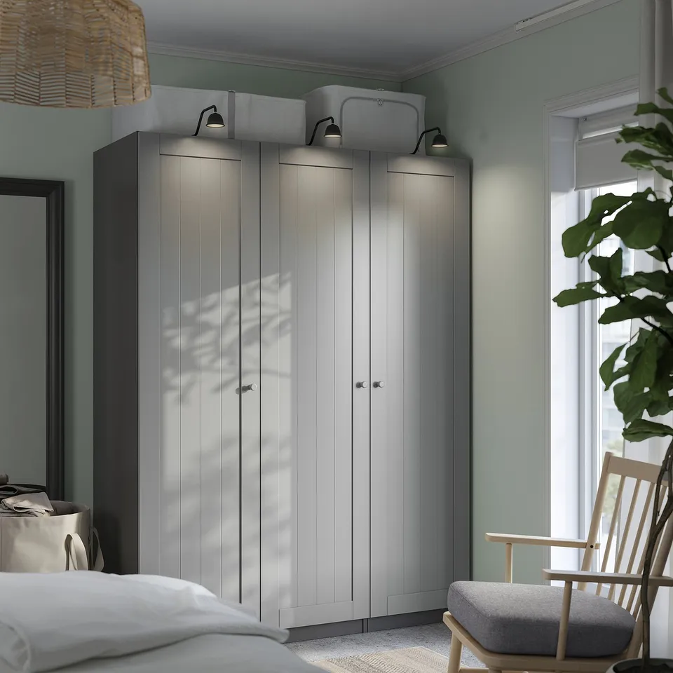 Minimalist bedroom with light grey PAX wardrobe, tall and wide, matching bed, small chair, and large plant. Soft lighting.