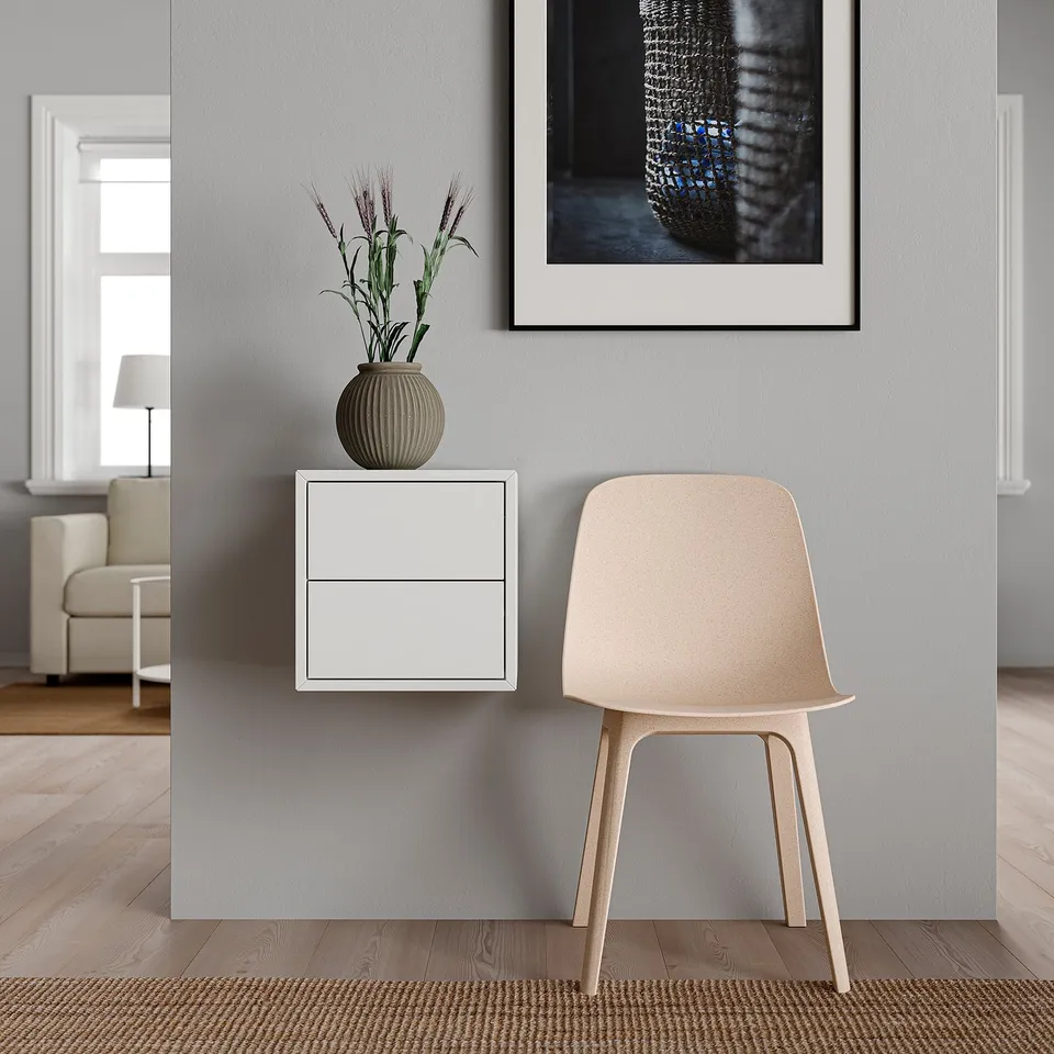 Minimalist room with wall-mounted white EKET cabinet, light wooden chair, and greyish rug. Decorated with a textured vase and framed art.