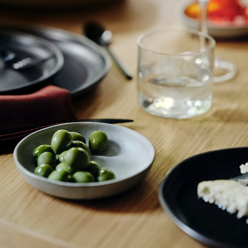 Small bowl of bright green peas on a grey plate, glass of clear water, and a small dish with white, crumbly food next to them on a wooden table.
