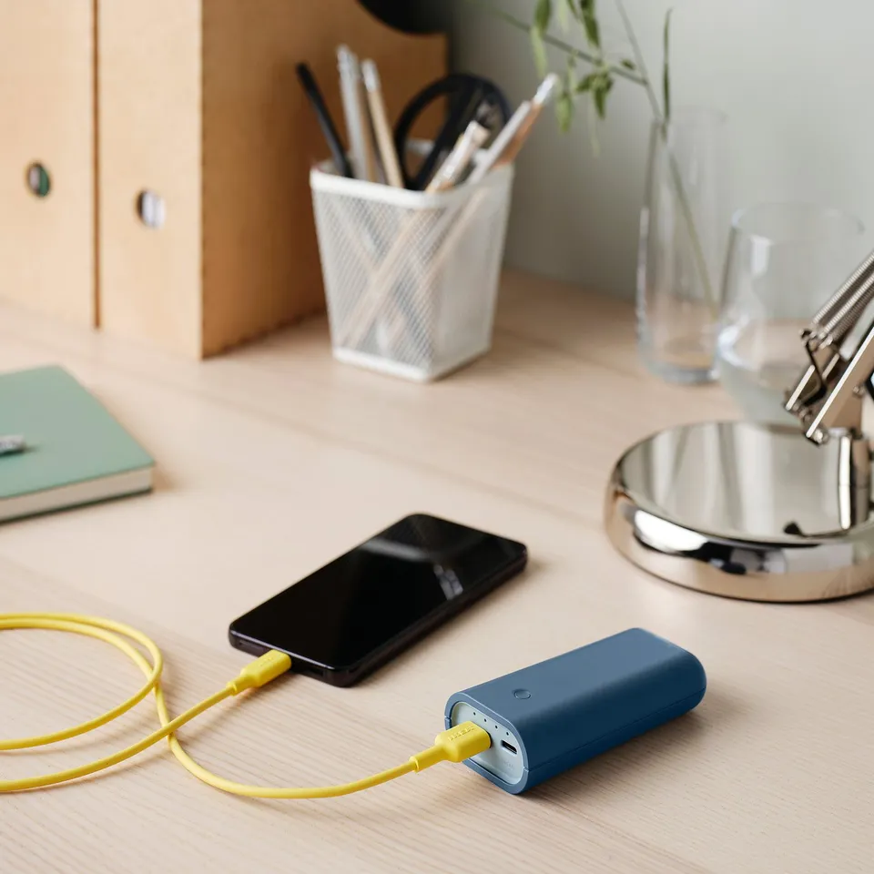 Desk with blue VARMFRONT power bank charging black smartphone, yellow cable. Nearby, pens, notebook, and lamp.