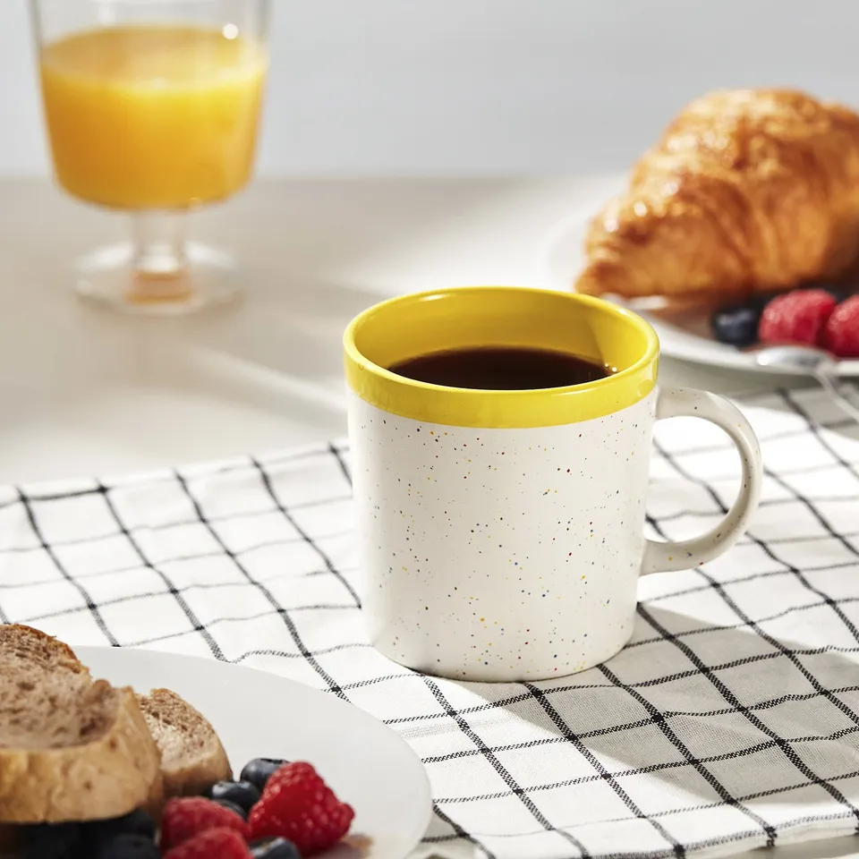 A breakfast table with a yellow-rimmed mug filled with coffee, next to a glass of orange juice, croissants, and berries.