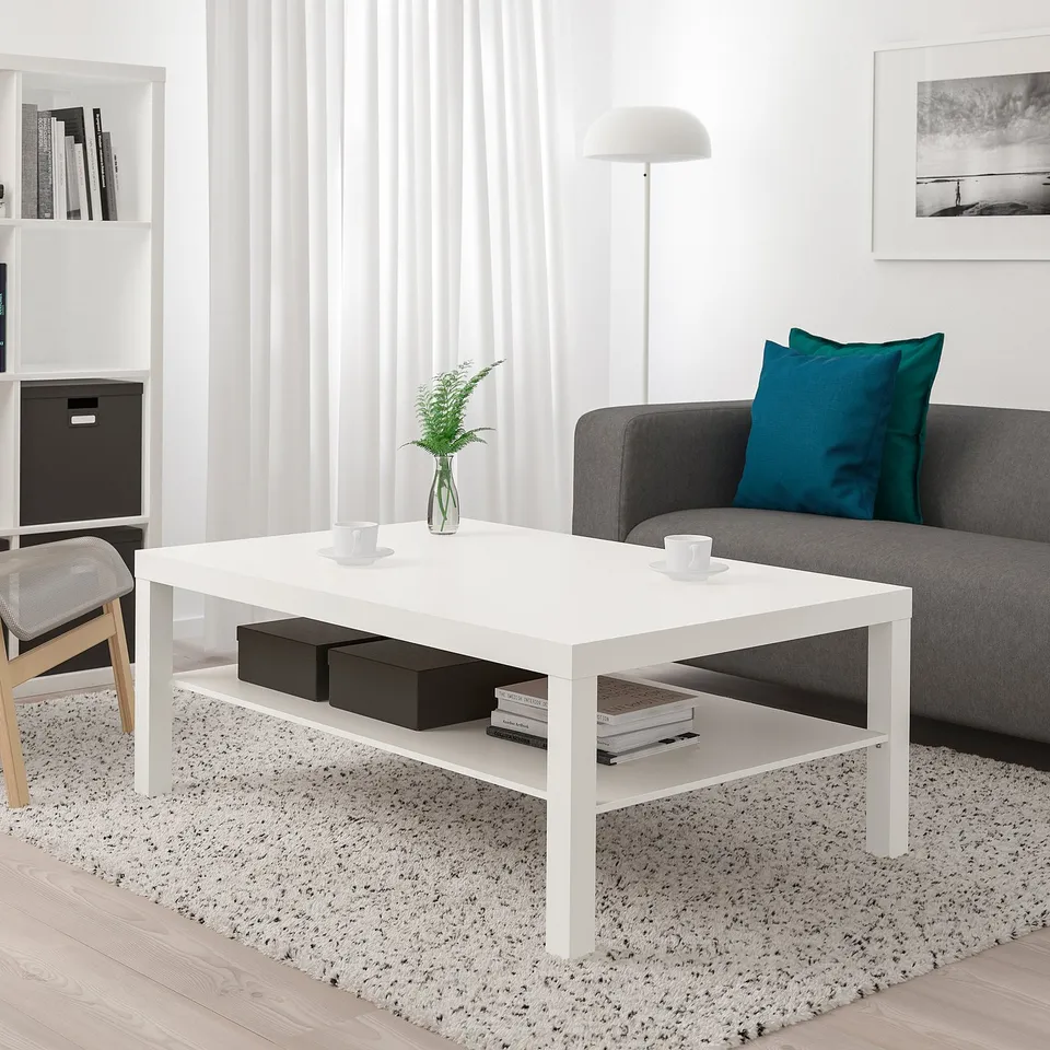 Modern living room with LACK white coffee table at centre, grey sofa, white curtains, and bLACK storage unit. Table holds two cups and a plant.