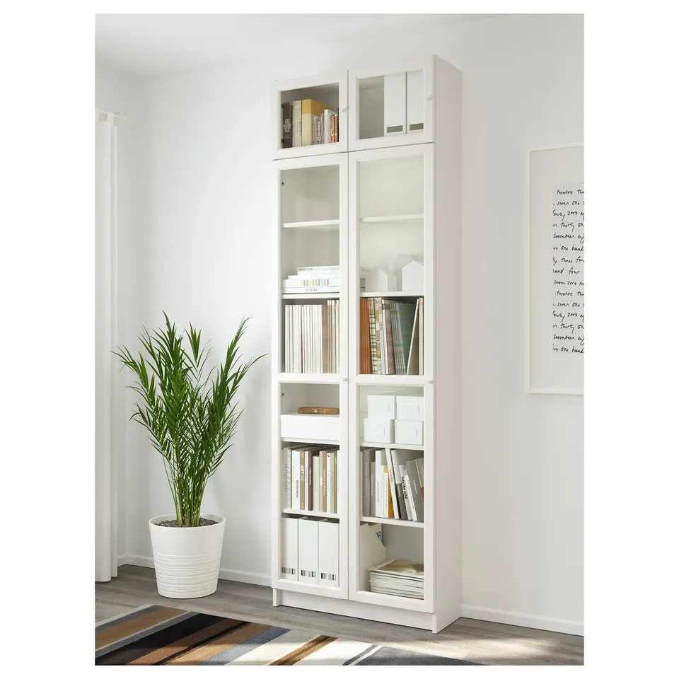 Bookshelf with glass doors, holding books and decor, adjustable shelves, against white wall, plant left.