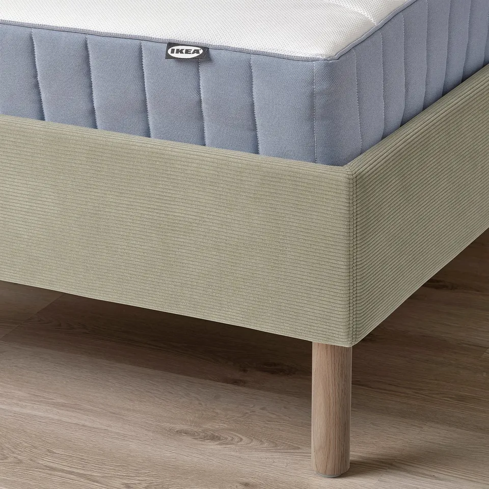 A corner of a TÄRNKULLEN bed frame with a light grey cover and a padded headboard, resting on wooden legs. The bed base is visible, with a IKEA tag.