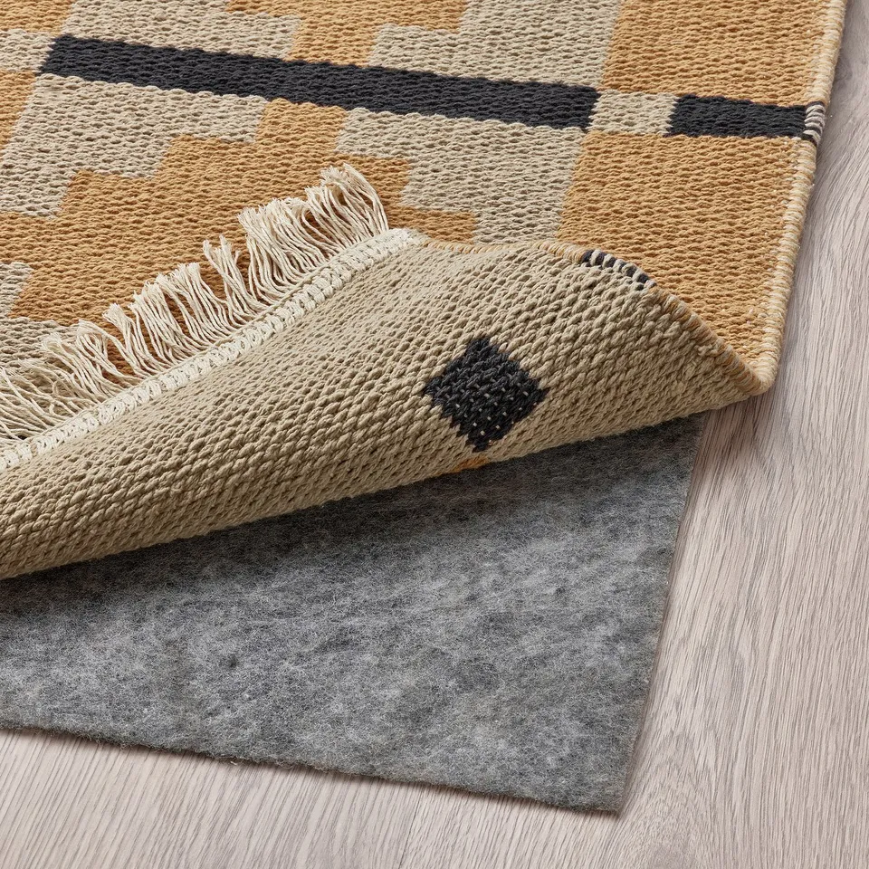 A beige and dark geometric rug is shown partially folded back on a wooden floor, revealing a grey non-slip backing.