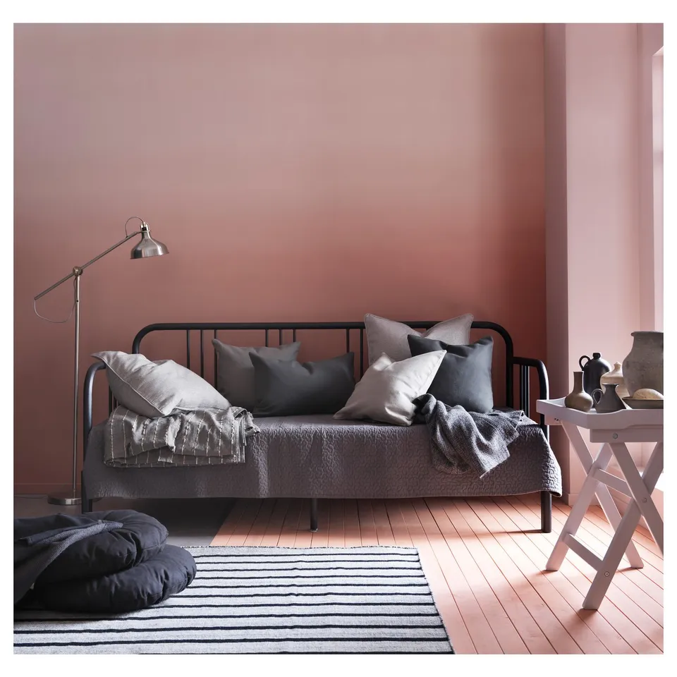 Modern, minimalist day-bed. Black metal frame, grey cushions, soft grey blanket. Stylish floor lamp. Cosy guest space.