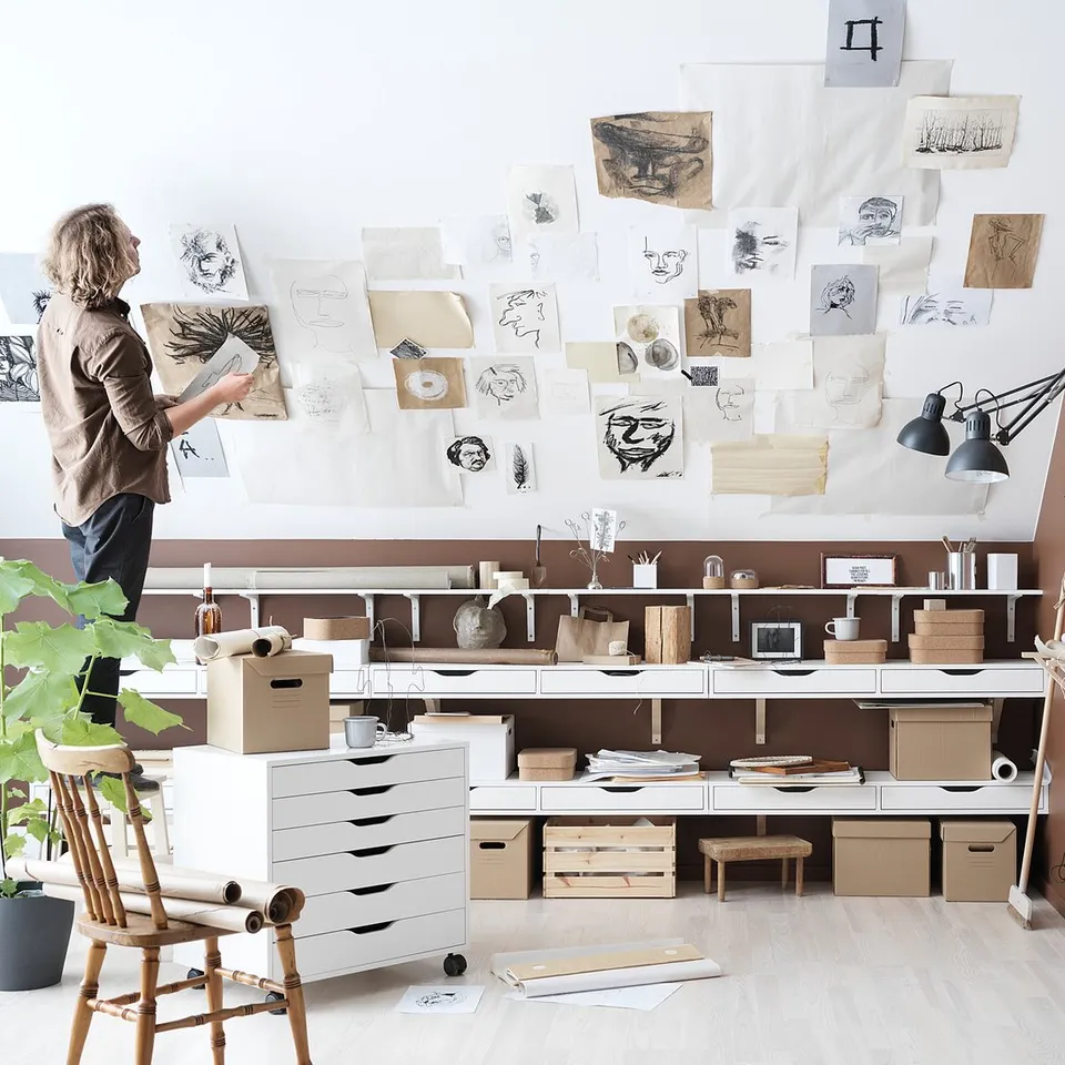 Art studio featuring ALEX drawers in white, various art supplies, sketches pinned on wall, wooden chair, lamp, and plant.