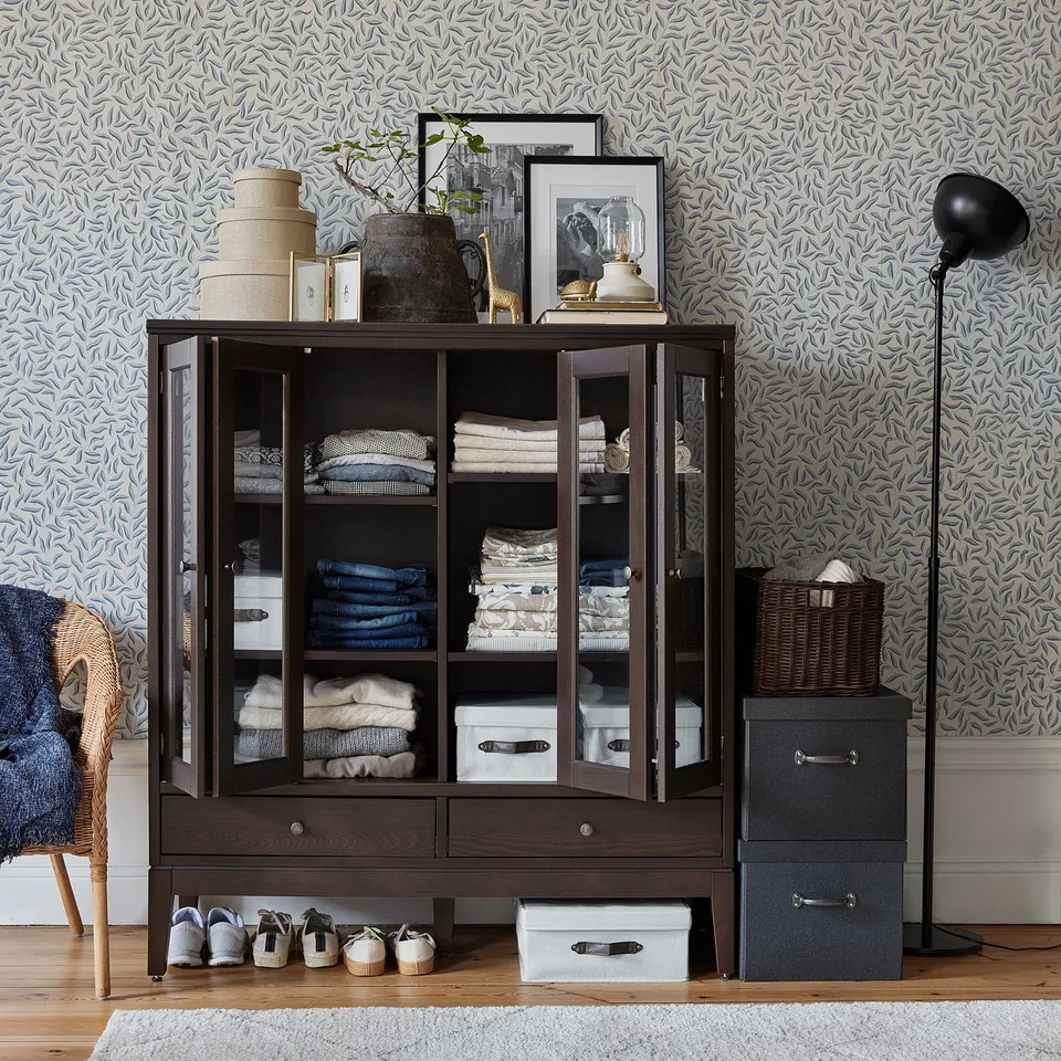 Glass-fronted wardrobe with clothes, vases, frames. Blue wallpaper, wicker basket, lamp.
