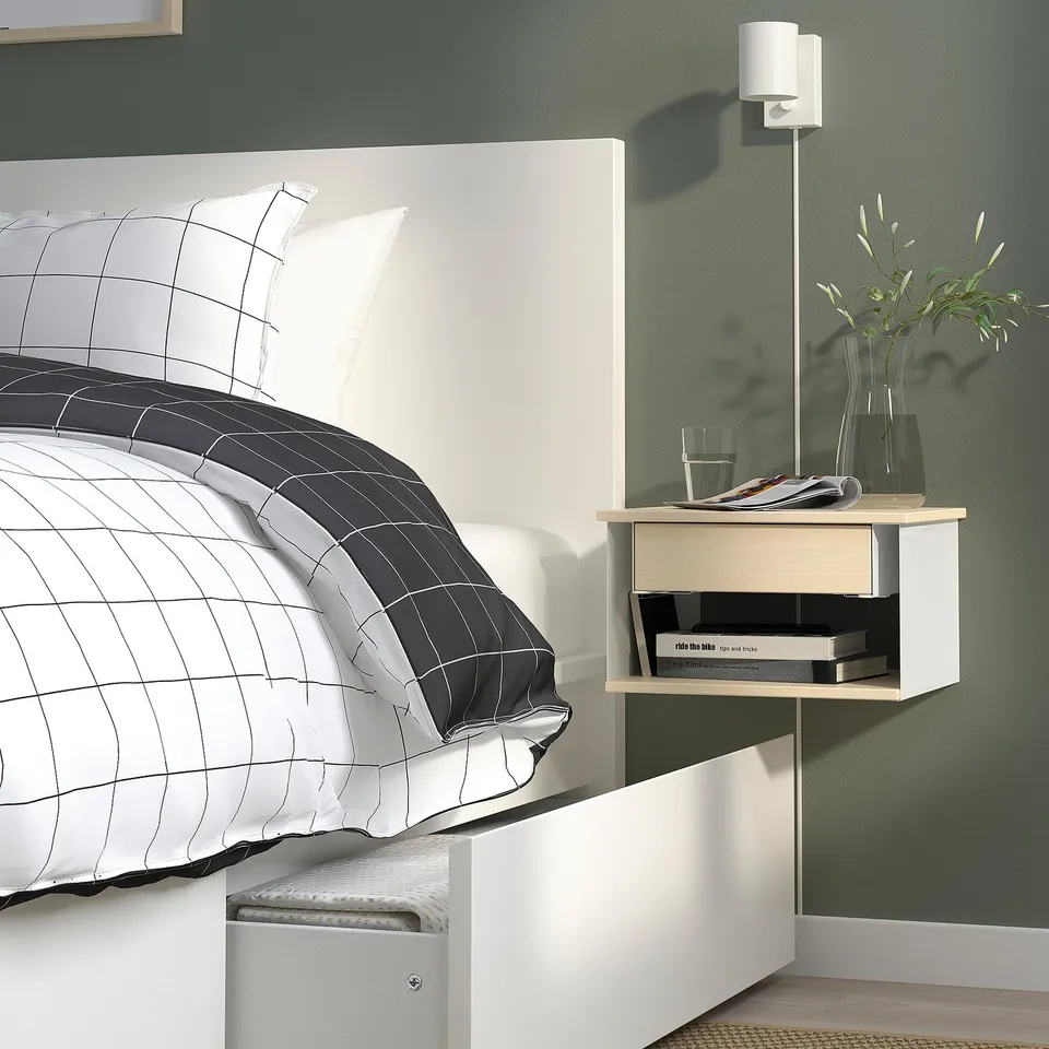 Bedside table with open storage, wall-mounted, holding books, a vase with plants, and a glass.