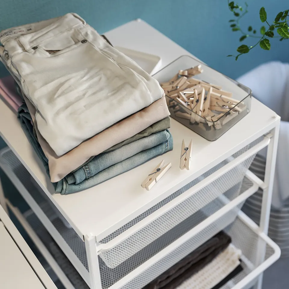 Folded clothes organised on white JONAXEL shelf with clothespins, placed against blue wall.