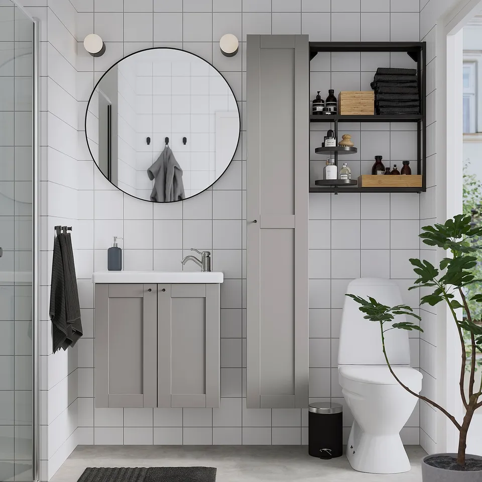 Modern bathroom with large round black-framed LINDBYN mirror above grey sink cabinet, white toilet, black accents, and wall shelves.