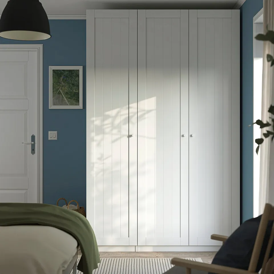 Modern bedroom with large white PAX wardrobe against blue wall, alongside bed, featuring clean lines and plenty of storage space.