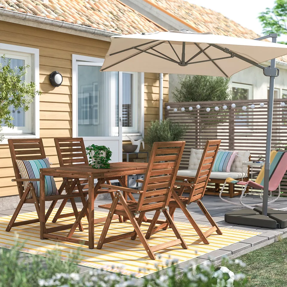Wooden patio table & chairs on striped rug, with umbrella & cushioned seating in background, backyard with house & plants.