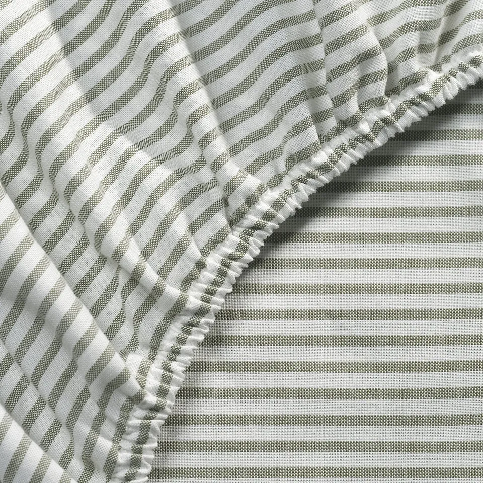 Close-up of textured, striped bed sheet. Visible elastic hem, indicating quality and fit. White and grey stripes, made of 100% cotton.