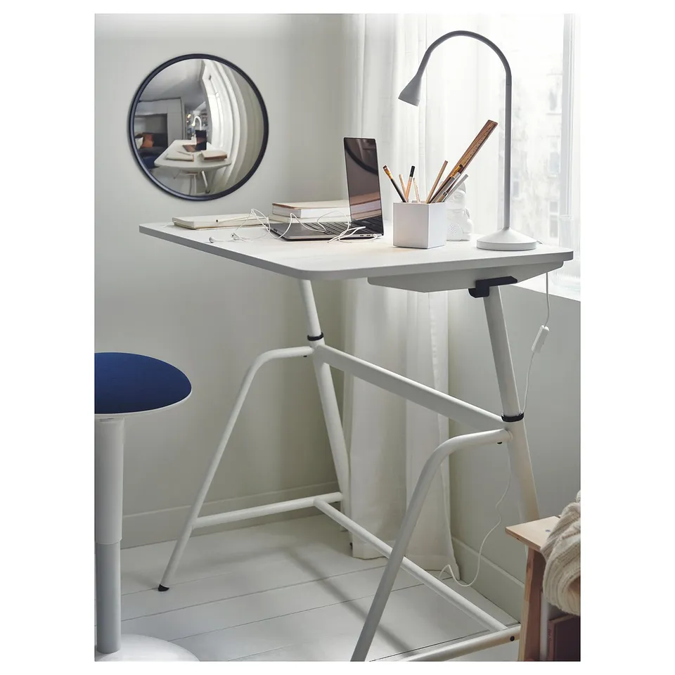 A small, white, modern desk setup. It includes a laptop, desk lamp, and mirror. The mirror is round, and the desk has a unique triangular metal base.