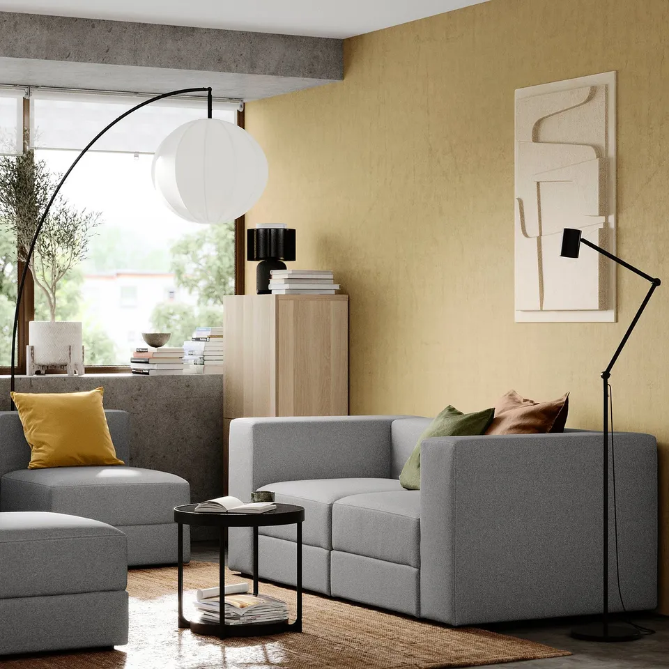 Modern living room with grey JÄTTEBO modular sofa, yellow pillows, floor lamp, side table, bookshelf with books, and abstract wall art.