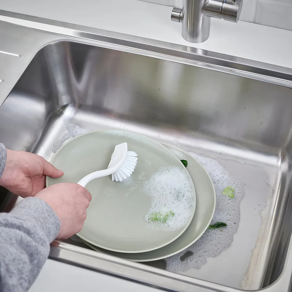Person washing dishes in sink using brush, with scraper edge for tough food debris, and hole in handle for hanging.