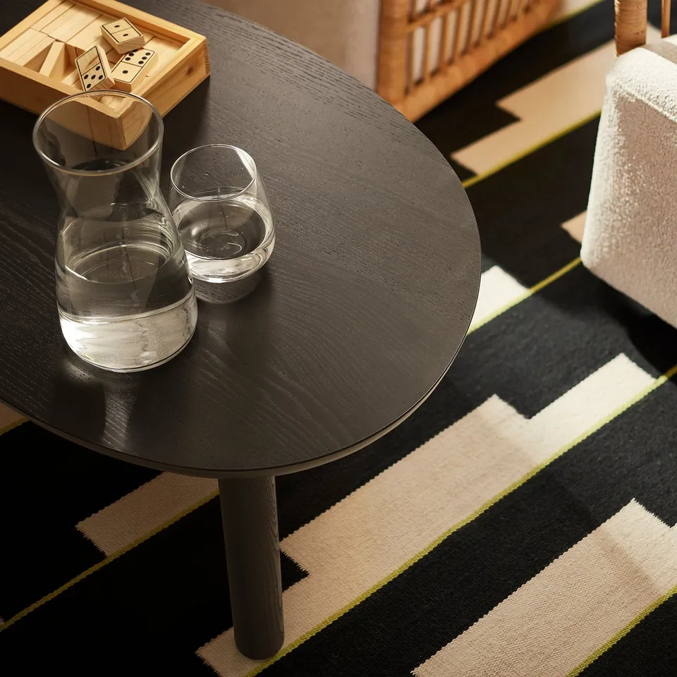 Top view of round ÖRTRÄSK coffee table with water glass and pitcher. Its small, black, wooden, and sits on a striped rug.