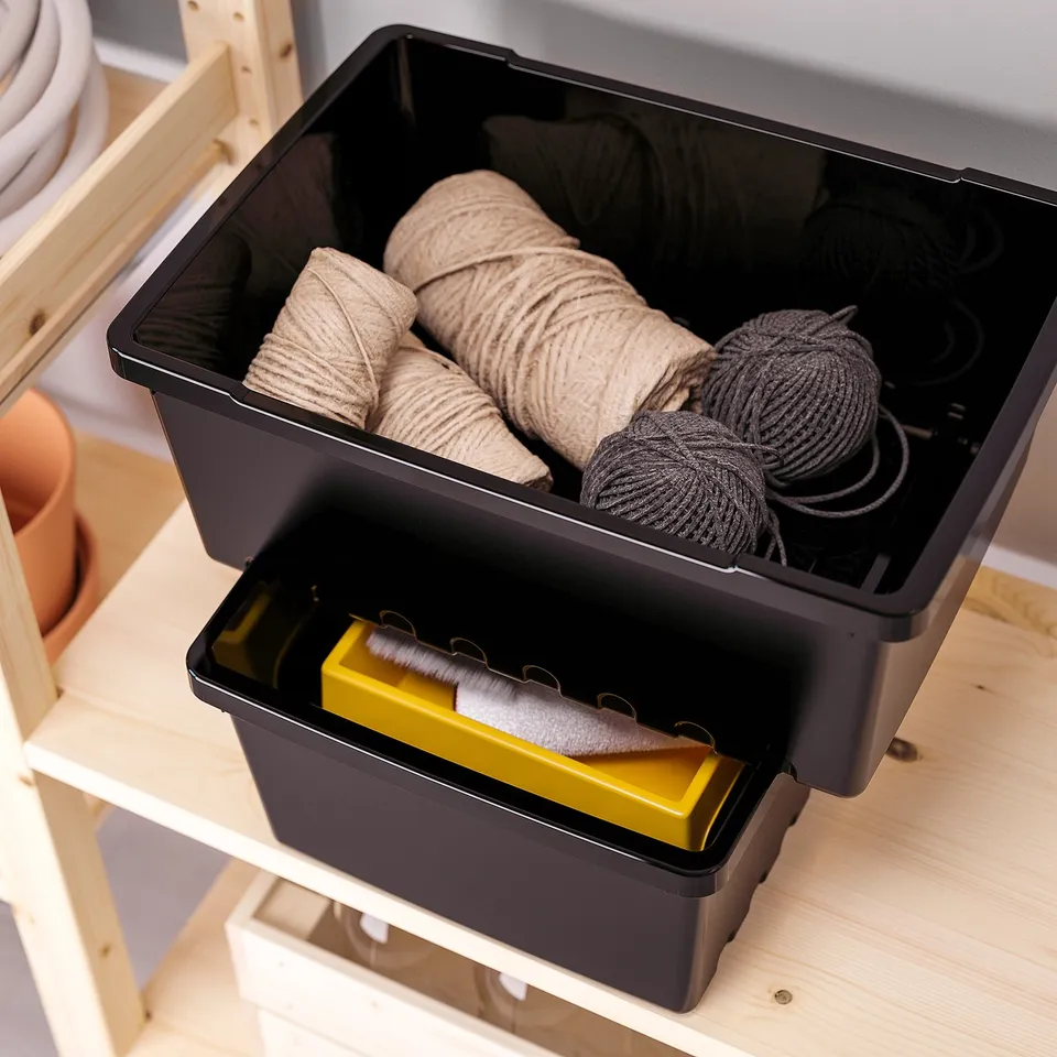 Plastic storage bin with two compartments, containing yarn and brushes, organised on wooden shelf.