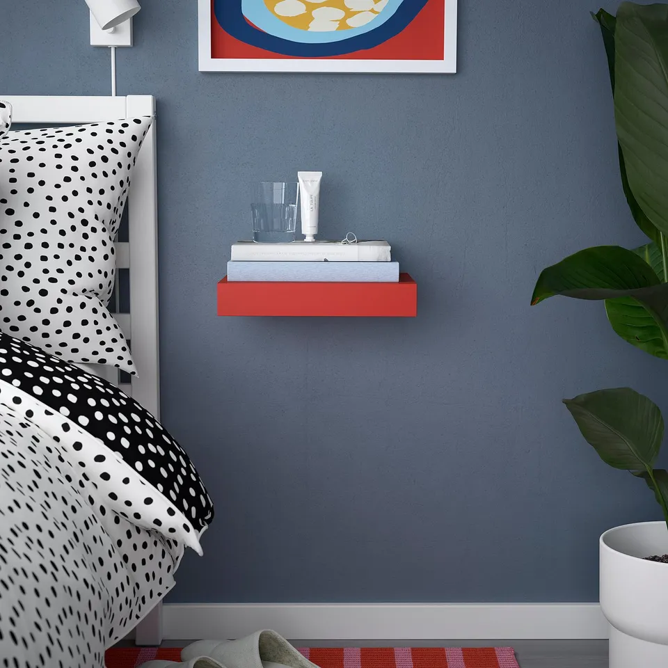 Red LACK wall shelf holds books and items, brightening space. Positioned near bed, below artwork, beside large plant.