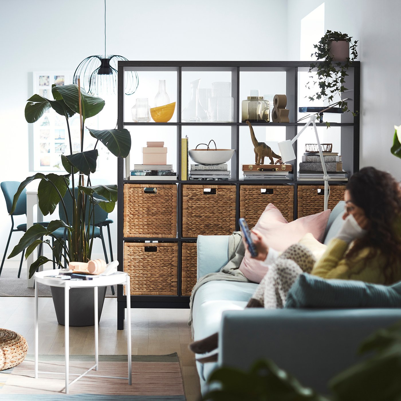 Modern living room with KALLAX shelf, blue sofa, plant, book, and person using smartphone.