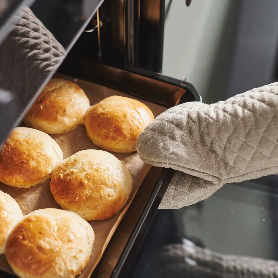 Hand with oven glove removing tray of freshly baked golden rolls from oven.