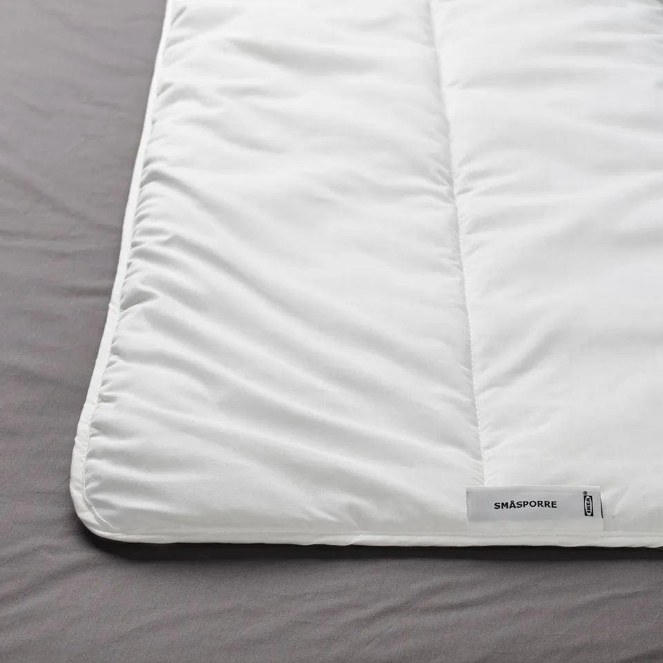 A close-up of a white duvet on a bed, showing its soft, plush texture and corner tag displaying SMÅSPORRE.