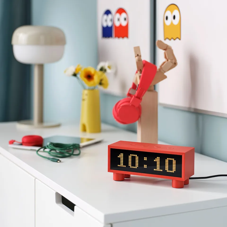 Red KORTLINJE clock on desk, showing time, temp, humidity. Surrounded by headphones, flowers, lamp.