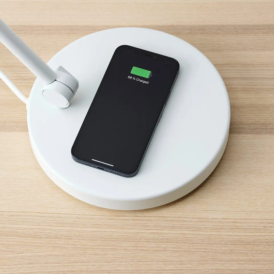 A smartphone wirelessly charging on a round, white NYMÅNE charging pad with a usb port and adjustable lamp.