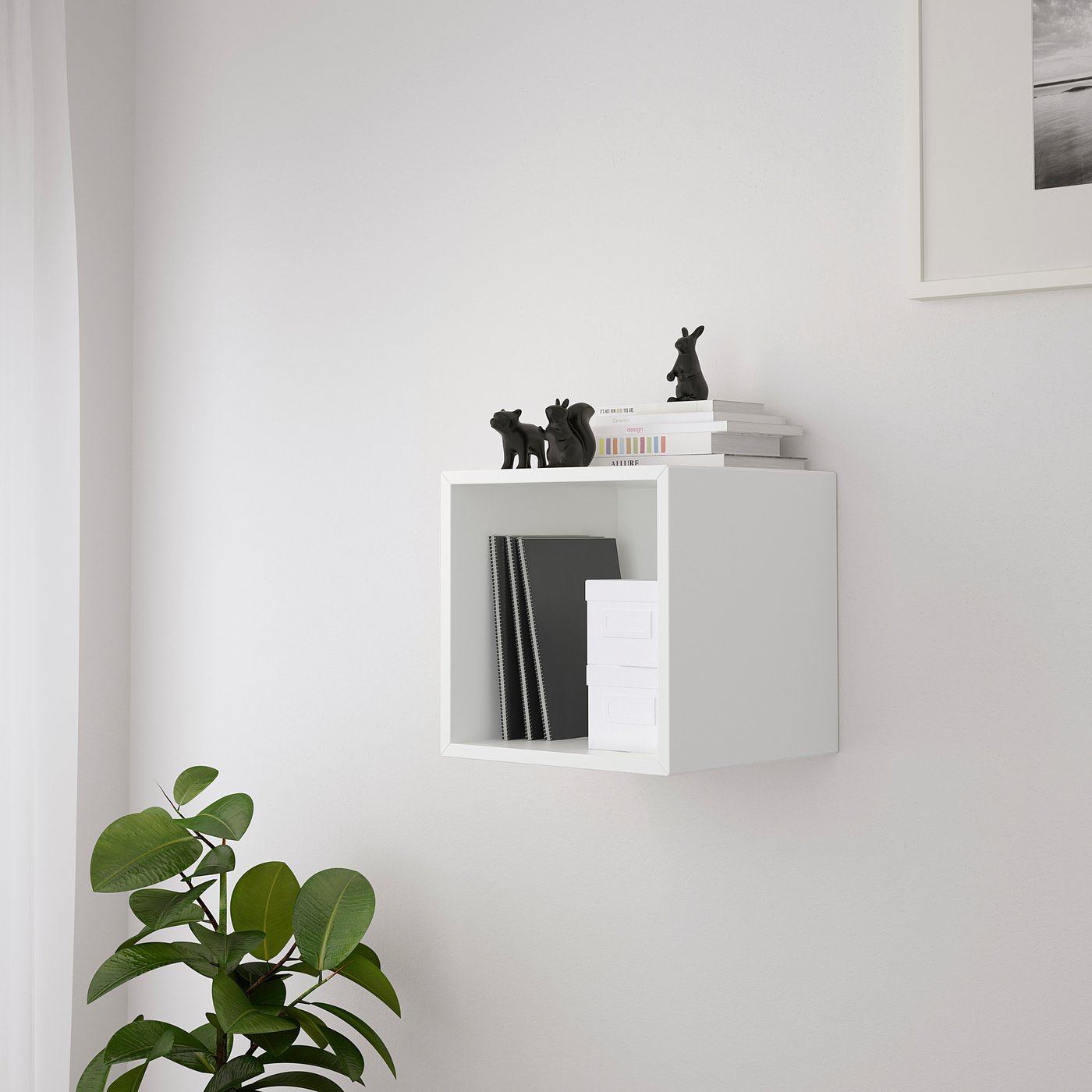 White wall-mounted cabinet EKET with open storage, holding various items including books, black figurines, and folders. Greenery at bottom left.