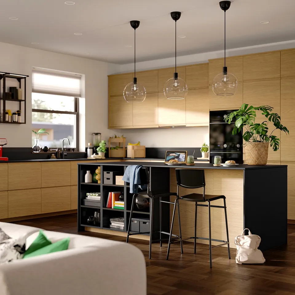 Modern kitchen with light wood cabinets, dark island, black stools, pendant lights, plant, and white sofa.