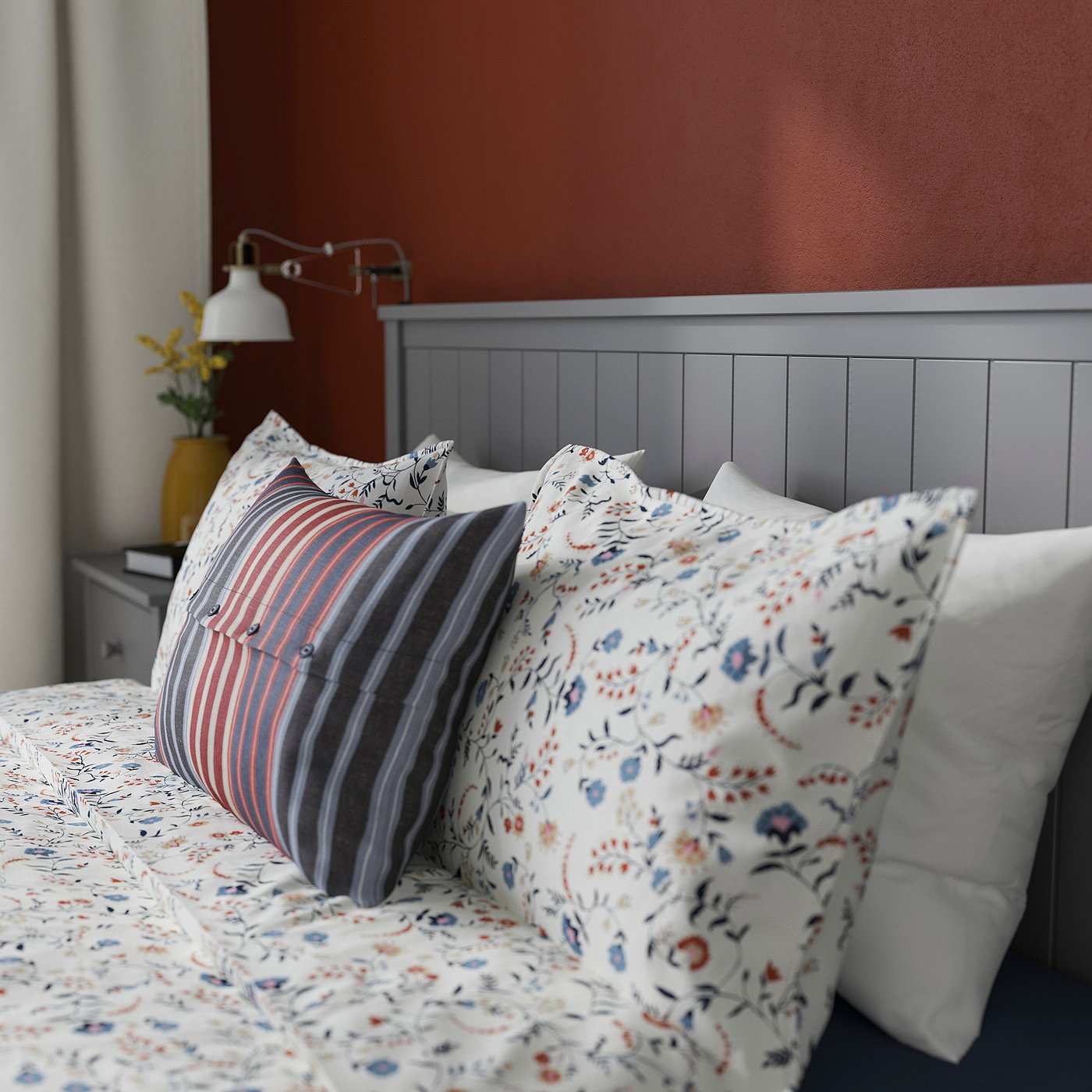Bed with floral pillows, striped cushion, grey headboard, wall light, and plant.
