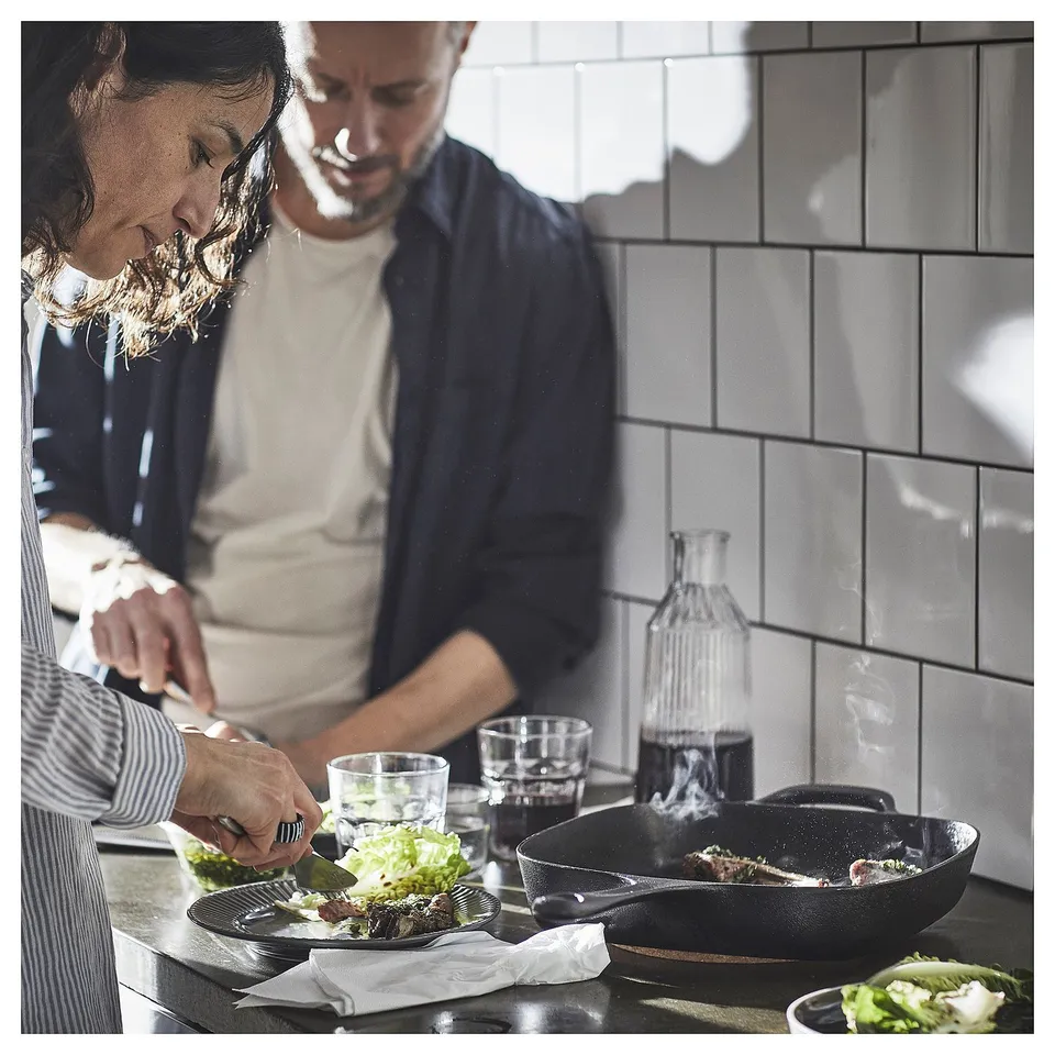 Two people cooking in kitchen. One stirs pan, other preps veggies. Modern, clean design.