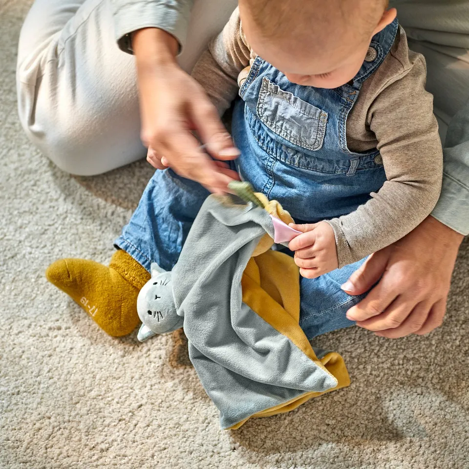 Baby playing with grey and yellow toy, wearing overalls, held by adult.