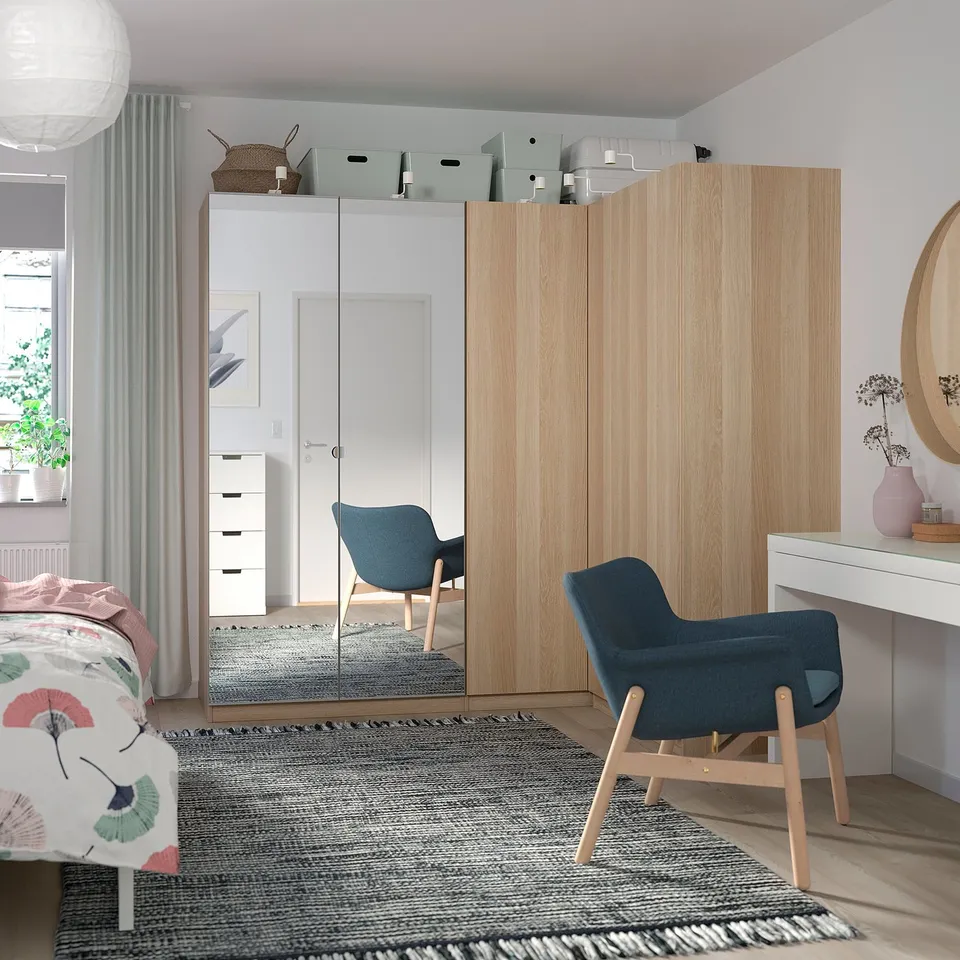 Modern bedroom with light wood pax wardrobe, blue chair, colourful bed, white drawer, round mirror.