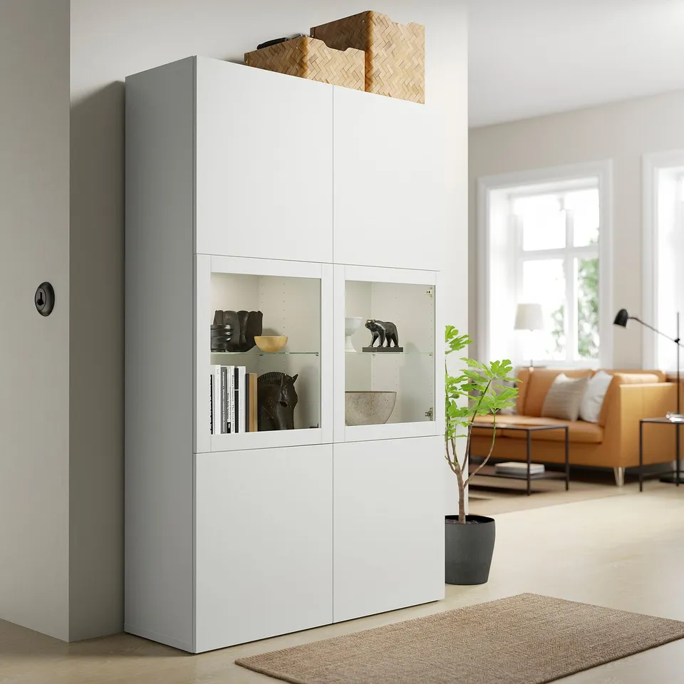 A white BESTÅ storage unit with glass doors. It holds decorative items and has a tall, rectangular shape. A plant and a cosy seating area are nearby.